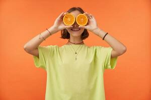 Porträt von attraktiv gegen Orange Hintergrund, nett suchen Mädchen mit kurz Brünette Haar halten Orangen Über ihr Augen, lächeln. Obst Stimmung. tragen Grün T-Shirt, Halskette, Armbänder und Ringe foto