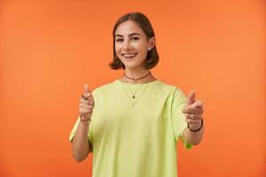 weiblich Student, attraktiv jung Dame mit kurz Brünette Haar lächelnd und zeigen zu das Kamera Über Orange Hintergrund. Sie habe Es. tragen Grün T-Shirt, Zähne Hosenträger und Armbänder foto