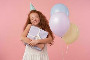 angenehm suchen Rothaarige wenig Mädchen mit lange lockig Haar drückt aus wahr positiv Emotionen, feiert etwas, isoliert Über Rosa Hintergrund im festlich Kleider und Geburtstag Deckel foto