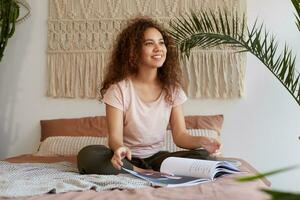 jung afrikanisch amerikanisch Frau mit lockig Haar, sitzt auf das Bett und verträumt sieht aus weg, lächelt und liest ein Neu Reise Zeitschrift, repräsentiert bevorstehende Reisen. foto