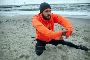 Porträt von aktiv jung dunkel behaart Mann mit Bart Hören zu Musik- mit Kopfhörer während Dehnen seine Muskeln Vor Laufen Übung. Sport und gesund Lebensstil Konzept foto