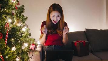 Junge Asiatin mit Tablet-Videoanruf im Gespräch mit Paar, Weihnachtsbaum mit Ornament auf dem Sofa im Wohnzimmer zu Hause geschmückt. soziale Distanzierung, Weihnachtsnacht und Neujahrsfest.