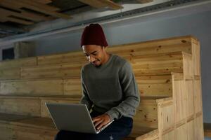 heiter gut aussehend attraktiv dunkel gehäutet bärtig Kerl im grau Pullover, Blau Hose und Burgund Deckel posieren Über Konferenz Zimmer, Sitzung Über hölzern Treppe mit modern Laptop auf seine Beine foto