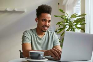 jung attraktiv afrikanisch amerikanisch Junge, sitzt beim ein Tabelle im ein Cafe, funktioniert beim ein Laptop, sieht aus beim das Monitor mit der Ekel, etwas falsch mit seine Liebling Seite? ˅. foto