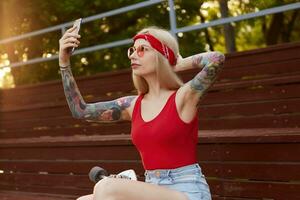 Porträt von jung schön blond Frau mit tätowiert Waffen im ein rot T-Shirt und Denim kurze Hose, mit ein gestrickt Bandana auf Kopf, im rot Gläser, halten Smartphone und macht Selfie zum ihr bloggen. foto