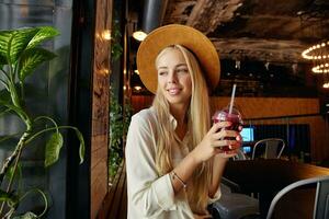 schön lange behaart blond Frau gekleidet im stilvoll Kleider Sitzung Über Restaurant Innere und suchen verträumt aus Fenster, halten Tasse mit trinken und Stroh im angehoben Hände foto