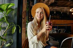 Innen- Foto von charmant ziemlich blond Dame tragen breit braun Hut und Weiß Hemd während Sitzung in der Nähe von Fenster im Stadt Cafe, suchen beiseite und lächelnd glücklich, haben Mittagessen brechen in der Nähe von Büro