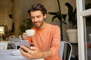 Innen- Schuss von gut aussehend jung Mann im pfirsichfarben Sweatshirt Trinken Kaffee im Stadt Cafe in der Nähe von Fenster, halten Telefon im Hand suchen beim Bildschirm foto