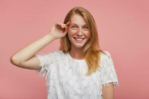 Studio Schuss von heiter schön jung weiblich mit foxy Haar berühren sanft ihr Gesicht mit angehoben Hand und suchen positiv beim Kamera mit breit lächeln, isoliert Über Rosa Hintergrund foto