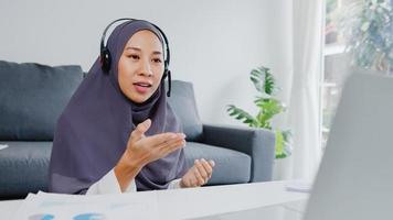 asiatische muslimische dame tragen kopfhörer mit laptop sprechen mit kollegen über den plan in konferenz-videoanrufen, während sie von zu hause im wohnzimmer arbeiten. soziale Distanzierung, Quarantäne zur Vorbeugung des Coronavirus. foto