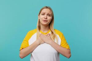 Süss jung ziemlich blauäugig lange behaart blond Frau halten Palmen auf ihr Truhe während suchen emotional beim Kamera, tragen beiläufig Kleider während posieren Über Blau Hintergrund foto