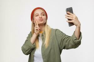 Porträt von jung schön lange behaart blond Frau schnüren ihr Lippen und geben zwinkern während Herstellung Foto von Sie selber und zeigen Sieg Geste, posieren Über Blau Hintergrund