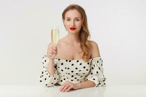 Porträt von jung schön blauäugig Frau mit lange blond Haar, mit rot Lippen im ein Tupfen Kleid, Standort beim das Tisch, halten ein Champagner Glas, isoliert Über Weiß Hintergrund. foto