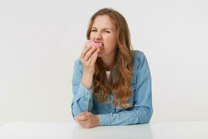 Porträt von ein jung ziemlich Frau tragen ein Denim Shirt, mit lange blond wellig Haar, fiel von das Diät und eifrig Essen ein Krapfen, isoliert Über Weiß Hintergrund. foto