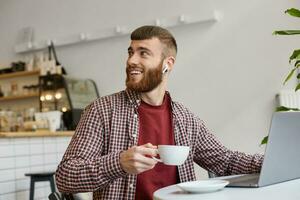 Foto von attraktiv Ingwer bärtig Mann Arbeiten beim ein Laptop während Sitzung im ein Cafe, Trinken Kaffee, tragen im Basic Kleidung, suchen zurück, Vielen Dank Barista zum ein wunderbar Kaffee.