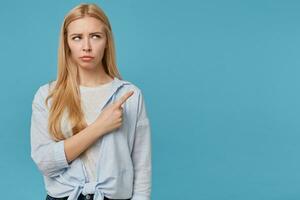 nachdenklich jung blond Dame mit lange Haar tragen Blau Hemd und grau T-Shirt, runzelte die Stirn mit gefaltet Lippen und zeigen beiseite mit Zeigefinger, isoliert Über Blau Hintergrund foto