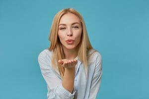 positiv jung blond weiblich mit beiläufig Frisur posieren Über Blau Hintergrund, erziehen Palme und weht Luft Kuss zu Kamera mit geschürzt Lippen, tragen Blau Hemd und grau T-Shirt foto