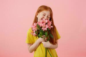 Porträt zierlich lächelt Sommersprossen rothaarig Mädchen mit zwei Schwänze, sieht aus Niedlich, trägt im Gelb T-Shirt, hält Strauß und steht Über Rosa Hintergrund, genießen das Geruch von Blumen. foto