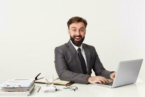 Porträt von ein Büro oben Manager Wer lächelt breit weil er gehört ein komisch scherzen, Sitzung beim Desktop im Büro, Arbeiten zum seine Laptop , gekleidet im ein teuer passen mit ein binden, genießen seine arbeiten. foto