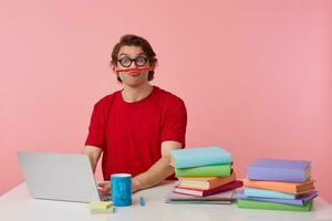 Porträt von komisch jung Schüler im Brille trägt im rot T-Shirt, sitzt durch das Tabelle und Arbeiten mit Laptop, sieht aus glücklich und froh, halten ein Bleistift mit seine Lippen isoliert Über Rosa Hintergrund. foto