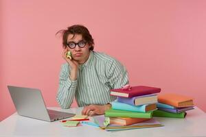 Studio Foto von schlagen jung männlich mit wild dunkel Haar Sitzung beim Arbeiten Tabelle Über Rosa Hintergrund, tragen Hemd und Brillen, behalten Kinn auf angehoben Hand und suchen zu Kamera müde