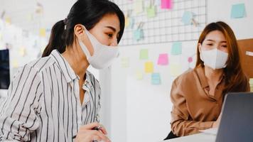 asiatische Geschäftsleute, die Computerpräsentationen verwenden, Brainstorming-Ideen über neue Projektkollegen und tragen eine schützende Gesichtsmaske im neuen normalen Büro. Lebensstil und Arbeit nach dem Coronavirus.