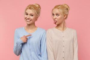 einer von schön jung blond Zwillinge ist täuschen um und zeigen ihr Zunge, ihr Schwester lächelt breit und zeigen beim ihr isoliert Über Rosa Hintergrund. foto