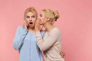 Foto von süß Zwillinge Blondinen, das Mädchen erzählt ihr Schwester das unglaublich Nachrichten um, das zweite einer geöffnet ihr Mund im Überraschung, steht Über Rosa Hintergrund.