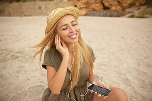 heiter jung lange behaart blond Dame behalten ihr Augen geschlossen während Hören zu Musik- und lächelnd glücklich, genießen Musik- während posieren Über Strand Hintergrund foto