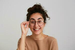 Porträt von heiter süß georgisch jung Frau mit lockig Haar trägt Beige zur Seite fahren und Brille fühlt sich glücklich und Lachen isoliert Über Weiß Hintergrund foto