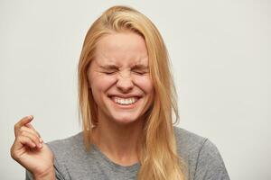 schließen oben von Lachen schön blond jung Frau, fühlt sich lustig, gehört ein Scherz oder komisch Geschichte, geschlossen ihr Augen, zahnig lächeln, trägt grau T-Shirt, isoliert Über Weiß Hintergrund foto