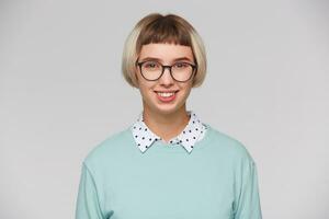 Nahansicht von heiter attraktiv jung Frau trägt Blau Sweatshirt und Brille fühlt sich Glücklich, lächelnd und sieht aus direkt im Kamera isoliert Über Weiß Hintergrund foto