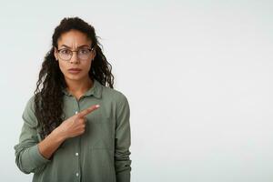 stark jung ziemlich dunkel gehäutet weiblich mit braun lockig Haar runzelte die Stirn Augenbrauen während zeigen Ernsthaft beiseite mit Index Finger, Stehen Über Weiß Hintergrund foto