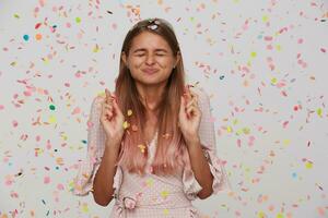 lächelnd süß jung Frau mit gekreuzt Finger und geschlossen Augen trägt Polka Punkt Rosa Kleid feiern Geburtstag und macht ein Wunsch isoliert Über Weiß Hintergrund mit Konfetti foto