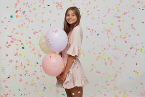 Porträt von glücklich attraktiv jung Frau mit lange gefärbt Pastell- Rosa Haar trägt Polka Punkt Rosa Kleid halten bunt Ballons im Hand und haben Party isoliert Über Weiß Hintergrund mit Konfetti foto