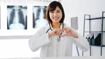 Selbstbewusste junge asiatische Ärztin in weißer medizinischer Uniform, die mit ihren Fingern eine Herzgeste macht und während der Videokonferenz mit dem Patienten im Krankenhaus lächelt. Beratungs- und Therapiekonzept.