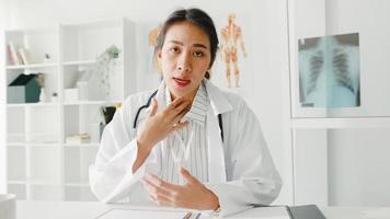 junge asiatische Ärztin in weißer medizinischer Uniform mit Stethoskop mit Computer-Laptop-Talk-Videokonferenz mit Patienten, Blick in die Kamera im Gesundheitskrankenhaus. Beratungs- und Therapiekonzept.