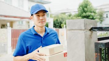 Junge asiatische Postzustellungskuriermann in blauem Hemd, das Paketkästen für den Versand an den Kunden im Haus handhabt, und asiatische Frauen erhalten ein geliefertes Paket im Freien. Paket einkaufen Lebensmittel Lieferkonzept. foto