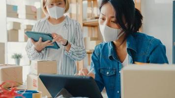 Junge Geschäftsfrauen in Asien tragen Gesichtsmaske Überprüfen Sie die Produktbestellung auf Lager und speichern Sie sie für die Arbeit mit Tablet-Computern im Home Office. Kleinunternehmer, Online-Marktlieferung, Lifestyle-Freelance-Konzept