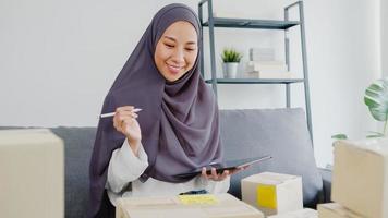Junge asiatische muslimische Geschäftsfrau Produktbestellung auf Lager überprüfen und auf Tablet-Computerarbeit im Home-Office speichern. Kleinunternehmer, Online-Marktlieferung, Lifestyle-Freelance-Konzept.