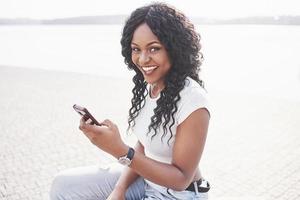 lächelndes schwarzes Mädchen mit einem Telefon am Wasser foto