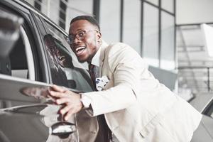 Schöner schwarzer Mann im Autohaus umarmt sein neues Auto und lächelt