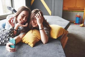 Zwei süße, lächelnde Zwillingsschwestern, die Smartphone halten und Selfie machen. Mädchen liegen auf der Couch posieren und Freude foto