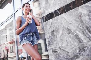 Porträt einer jungen stilvollen Hipster-Frau, die auf der Straße geht, ein süßes trendiges Outfit trägt, heißen Latte-Kaffee trinkt und lächelt