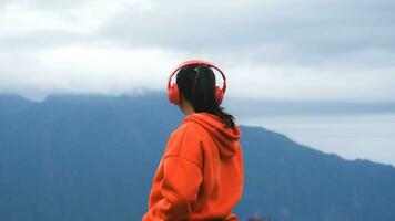 Rückansicht einer jungen Frau, die auf einem ruhigen Hügel steht und morgens Musik über Kopfhörer hört. frau, die einen pullover trägt, der die schönheit der natur genießt, die im winter den berg betrachtet. foto