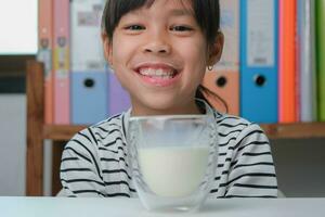 süß asiatisch Mädchen Trinken ein Glas von Milch beim Zuhause im Leben Zimmer. wenig Mädchen Trinken Milch im das Morgen Vor gehen zu Schule. gesund Essen im Kindheit. foto