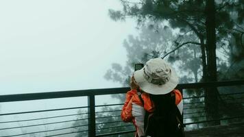 Rückseite Aussicht von ein jung Frau mit ein Rucksack Stehen auf ein Ruhe Berg oben und nehmen Bilder von das schön Berg Landschaft von das Morgen Nebel auf das Telefon. foto