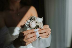 das Bräutigam Boutonniere ist vereinbart worden von Rose Blumen im das Hände von das Braut mit ein Französisch Maniküre im das wunderbar natürlich Licht von das Fenster foto