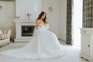 in voller Länge Porträt von das Braut im das Hotel Zimmer, das Mädchen ist Sitzung auf ein Stuhl im ein Hochzeit Kleid im das Mitte von das Zimmer mit ein Blume im ihr Hände foto