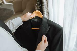 ein jung Mann hält ein Jacke im seine Hände im seine Zimmer, in der Nähe von das Fenster im ein Hotel Zimmer, Nahansicht Foto. das Bräutigam ist vorbereiten zum das Hochzeit Zeremonie foto